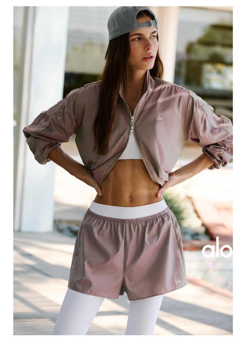 Model wearing mauve jacket, white top, shorts and cap standing outdoors for Alo