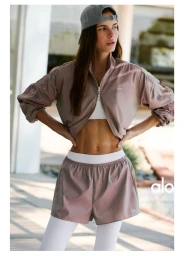Model wearing mauve jacket, white top, shorts and cap standing outdoors for Alo