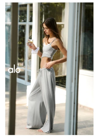 Model standing outdoors holding a glass of water, wearing a grey tank top and loose trousers for Alo.