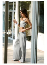 Model standing outdoors holding a glass of water, wearing a grey tank top and loose trousers for Alo.