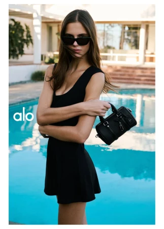 Model in a black dress and sunglasses holding a black handbag beside a swimming pool for Alo