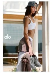 Model leaning against a pillar wearing a white sports bra, grey cap, and holding sneakers for Alo