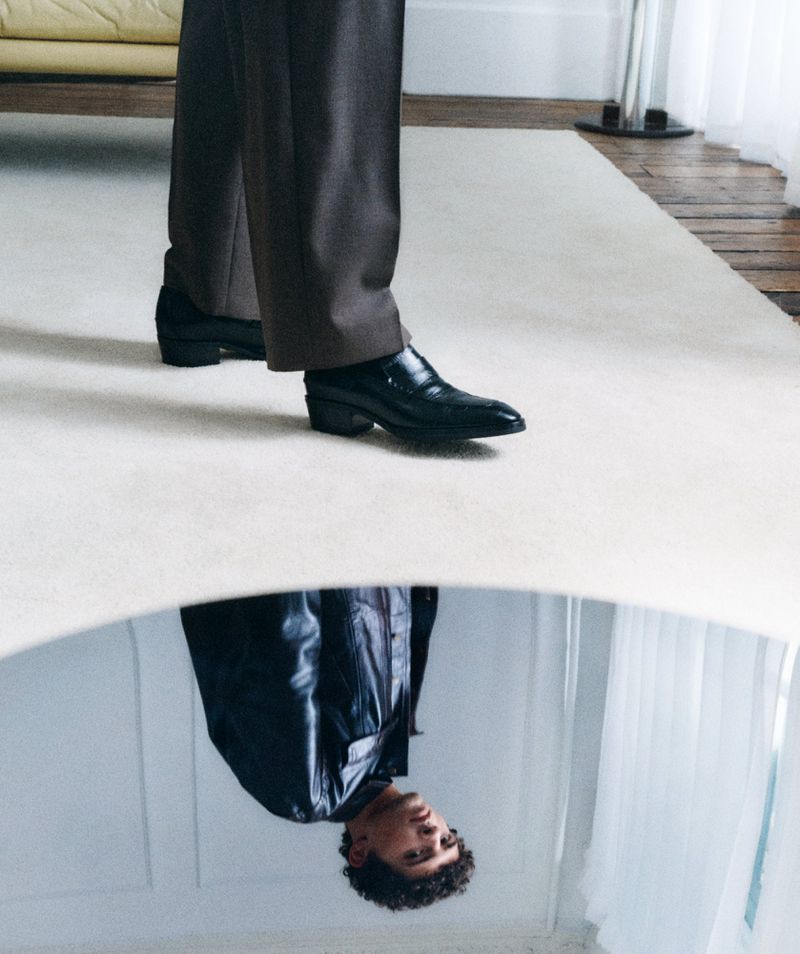 Model wearing black leather shoes and trousers reflected in a low curved mirror indoors