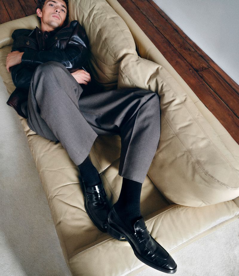 A male model reclining on a beige leather couch wearing a dark leather jacket, grey trousers, and black loafers.