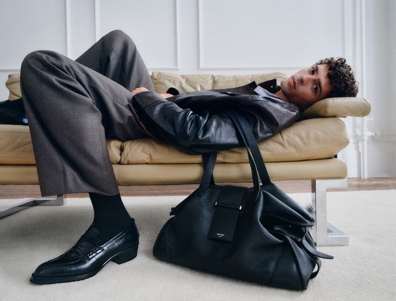 Model lounging on a beige leather daybed wearing a dark leather jacket, grey trousers, and black loafers beside a black leather duffel bag.