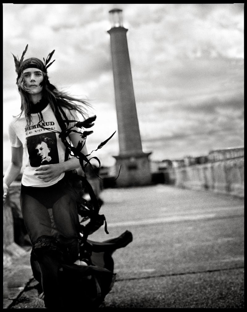 Model in feathered headband and graphic t-shirt walking outdoors near a tall tower in black and white