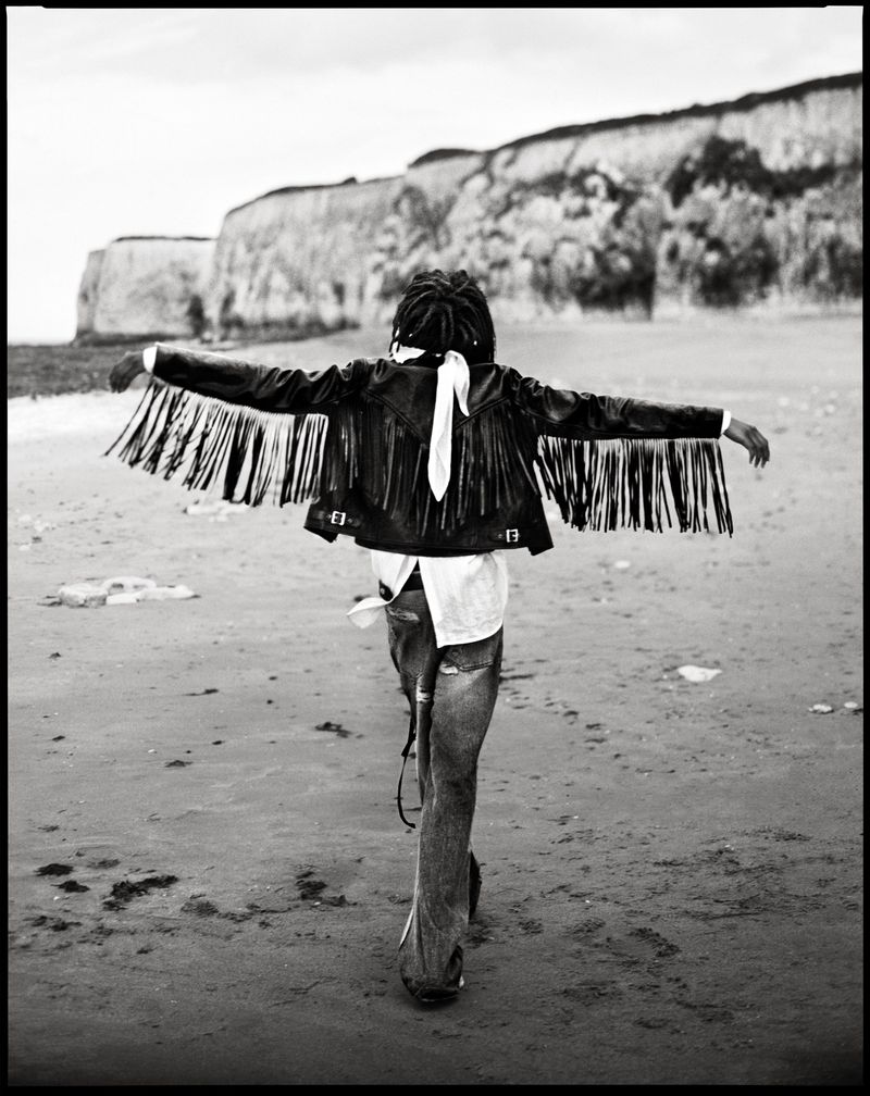 Model seen from behind wearing a fringed leather jacket on a sandy beach with cliffs