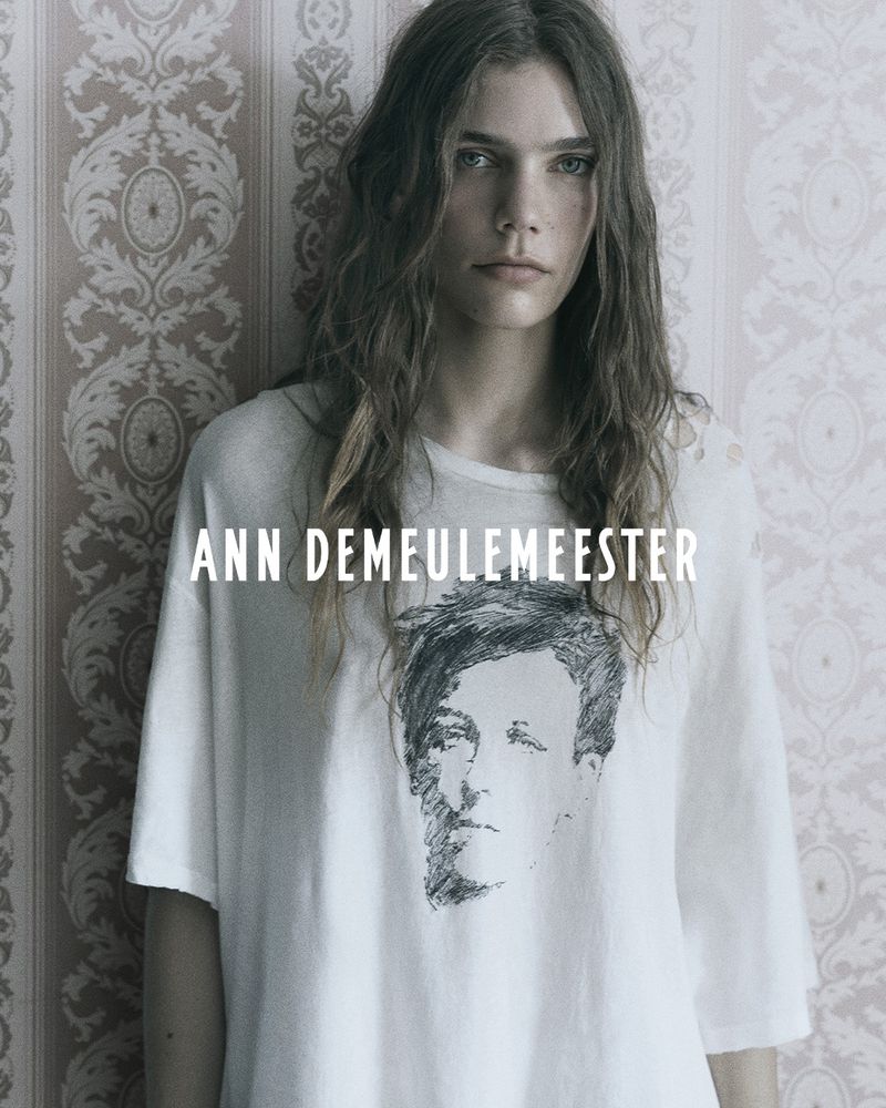 Model with long dark hair wearing a white graphic t-shirt against patterned vintage wallpaper.