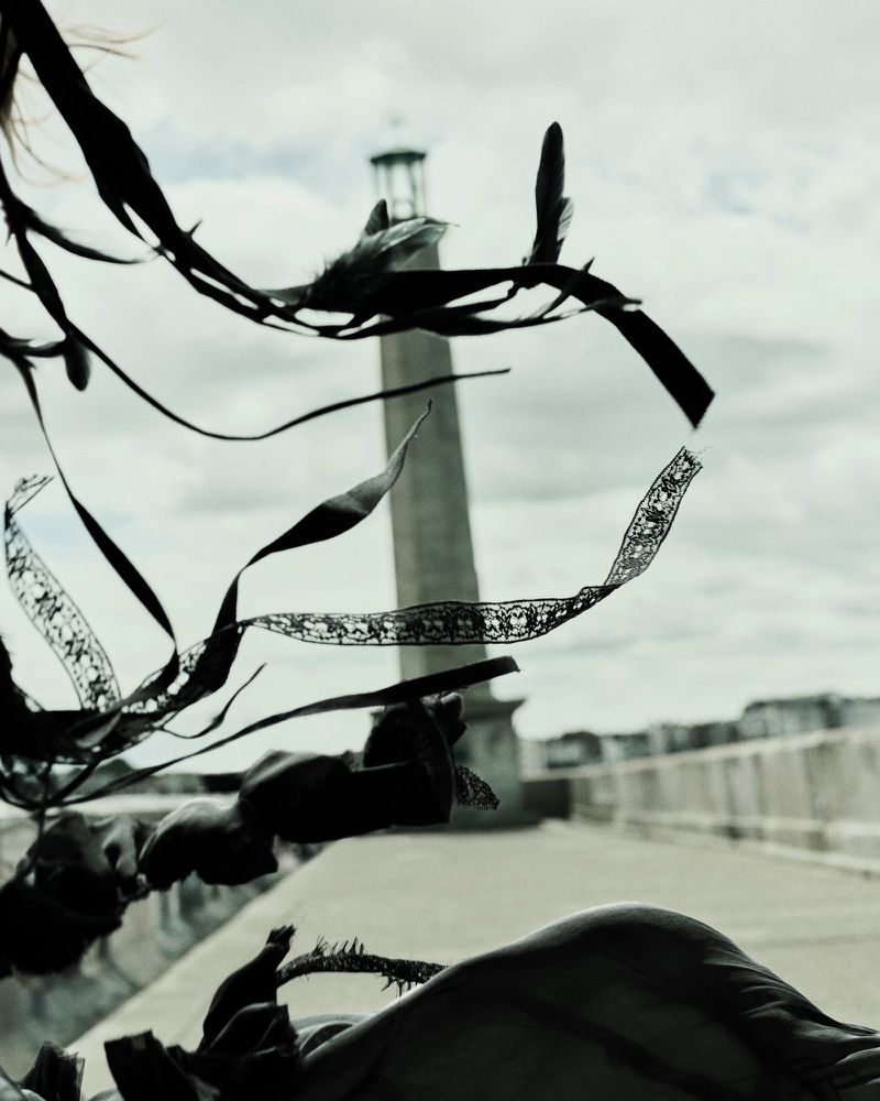A tall stone monument stands in the distance framed by dark flowing ribbons and lace elements in the foreground under an overcast sky.