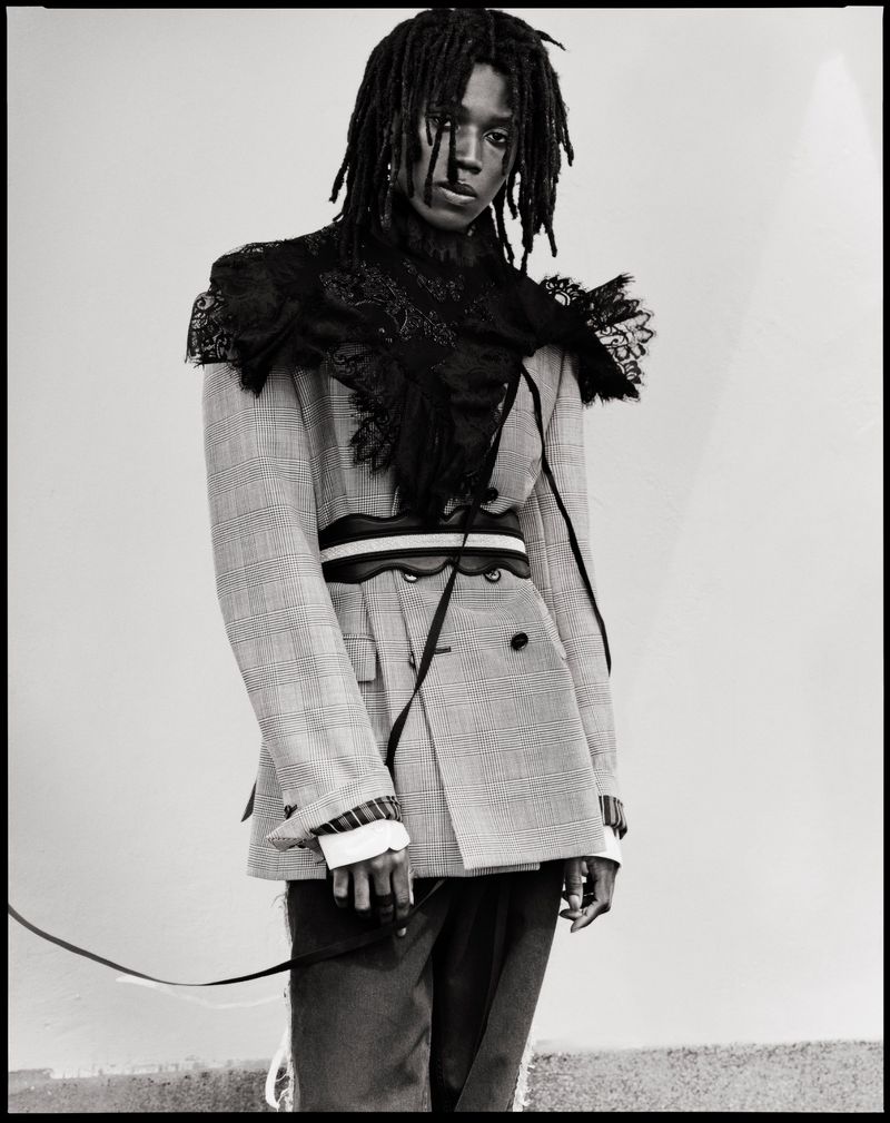 Model with dreadlocks wearing a patterned blazer with black lace collar and belt for Ann Demeulemeester