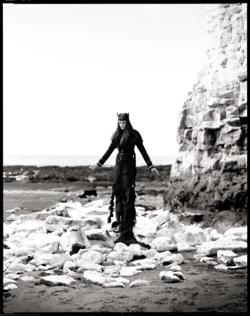 Model standing on a rocky beach wearing a dark outfit with arms outstretched next to a cliff