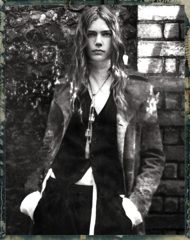 Monochrome portrait of a model with long hair wearing a dark vest and coat outdoors against a textured brick wall