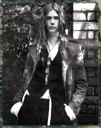 Monochrome portrait of a model with long hair wearing a dark vest and coat outdoors against a textured brick wall