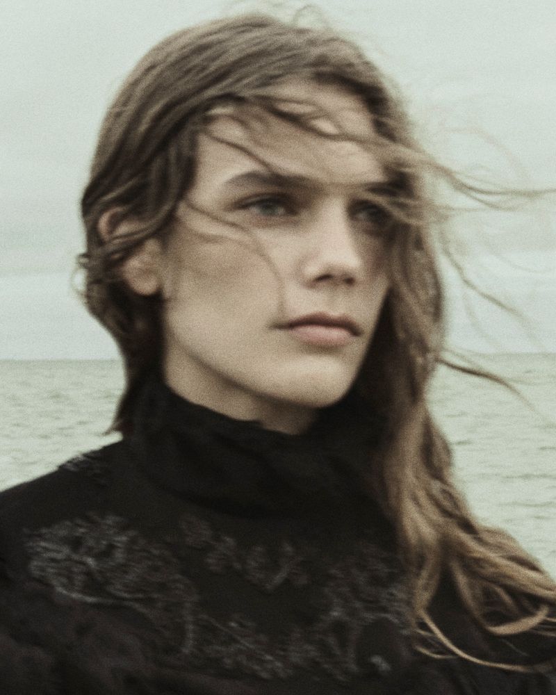 Close-up portrait of a model with long hair blowing in the wind wearing a dark embroidered garment by the ocean.