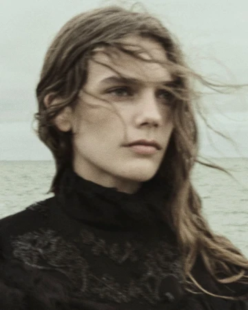 Close-up portrait of a model with long hair blowing in the wind wearing a dark embroidered garment by the ocean.