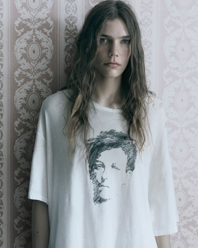 Model with long wavy hair wearing an oversized graphic t-shirt against a patterned vintage wallpaper background