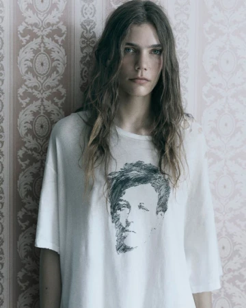 Model with long wavy hair wearing an oversized graphic t-shirt against a patterned vintage wallpaper background