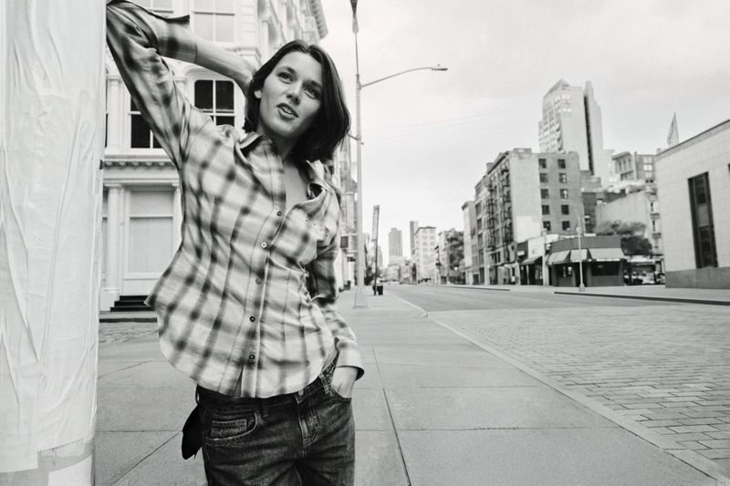 Black and white editorial image of a model wearing a plaid shirt and jeans, posing on a city street.