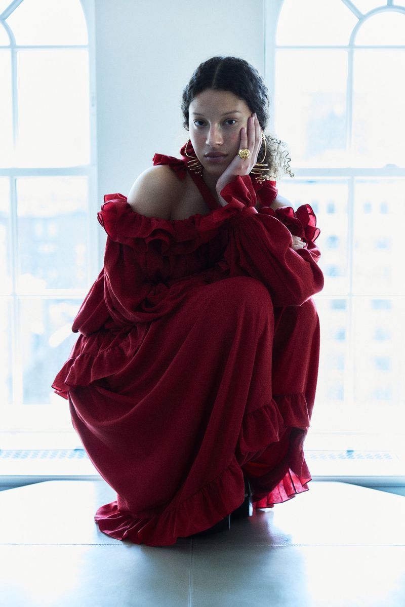 Model in a red ruffled dress crouching against tall arched windows in a light-filled interior