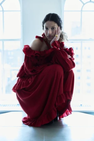 Model in a red ruffled dress crouching against tall arched windows in a light-filled interior