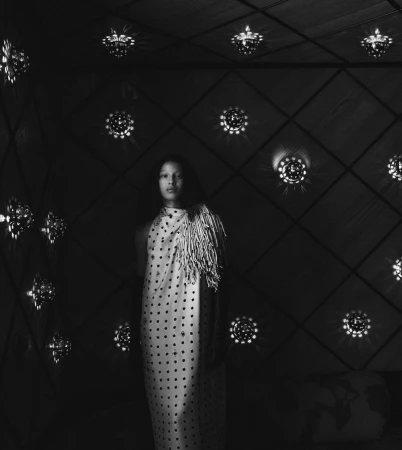 Model in a patterned sleeveless dress standing against a dark wall punctuated by glowing circular lights