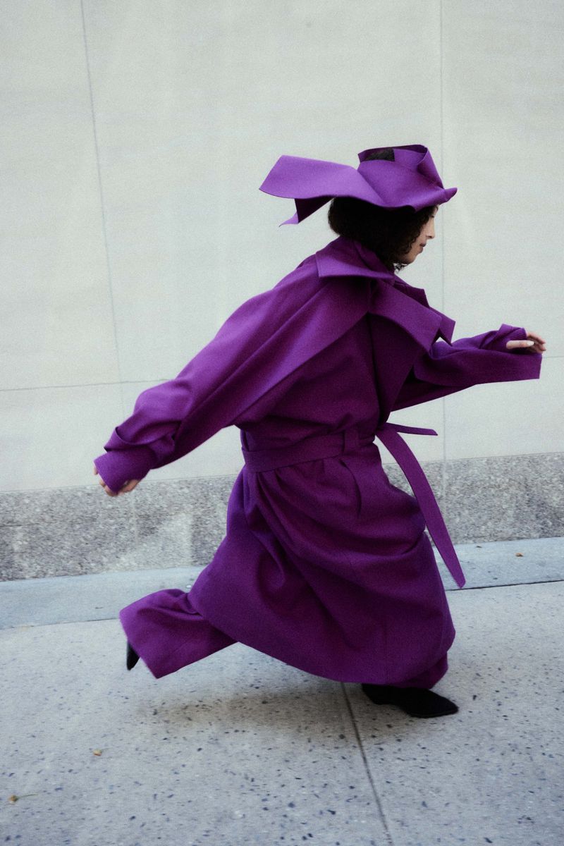 Model in a structured purple coat and matching hat walking along a sidewalk for Cultured Magazine.