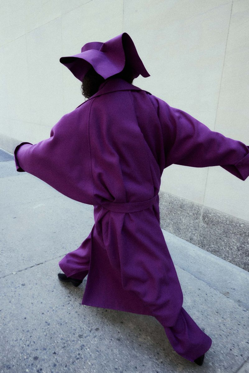 Model in a structured purple coat and matching hat walking down a stone sidewalk.