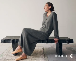 Woman in grey knit cardigan and long skirt sitting on a dark wooden bench wearing tan loafers against a plain wall