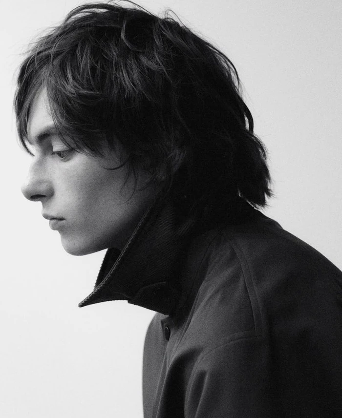 Monochrome portrait profile of a young man with dark medium-length hair wearing a dark high-collared jacket against a white background.