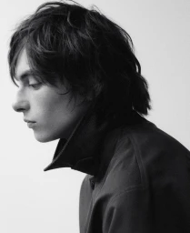 Monochrome portrait profile of a young man with dark medium-length hair wearing a dark high-collared jacket against a white background.