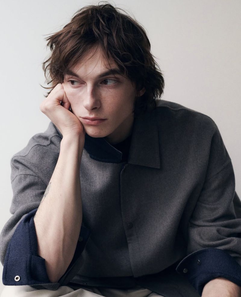 Close-up portrait of a young man with dark hair resting his chin on his hand, wearing a grey collared shirt against a white wall.