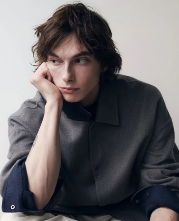 Close-up portrait of a young man with dark hair resting his chin on his hand, wearing a grey collared shirt against a white wall.