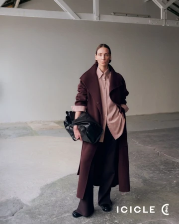 Model wearing burgundy coat and pink shirt holding a black bag standing in an industrial space with white walls.