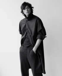 Monochrome portrait of a male model wearing a dark oversized coat and matching trousers against a plain white background.