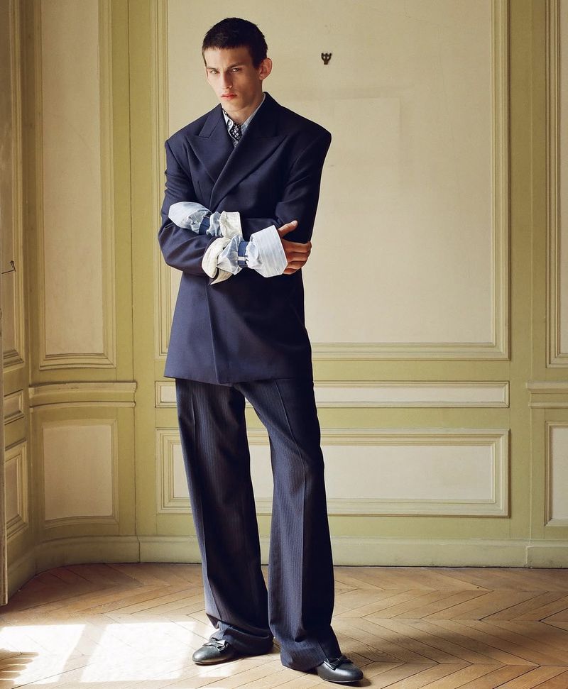 Model in an oversized navy suit with broad shoulders standing inside a room with classic wall paneling.