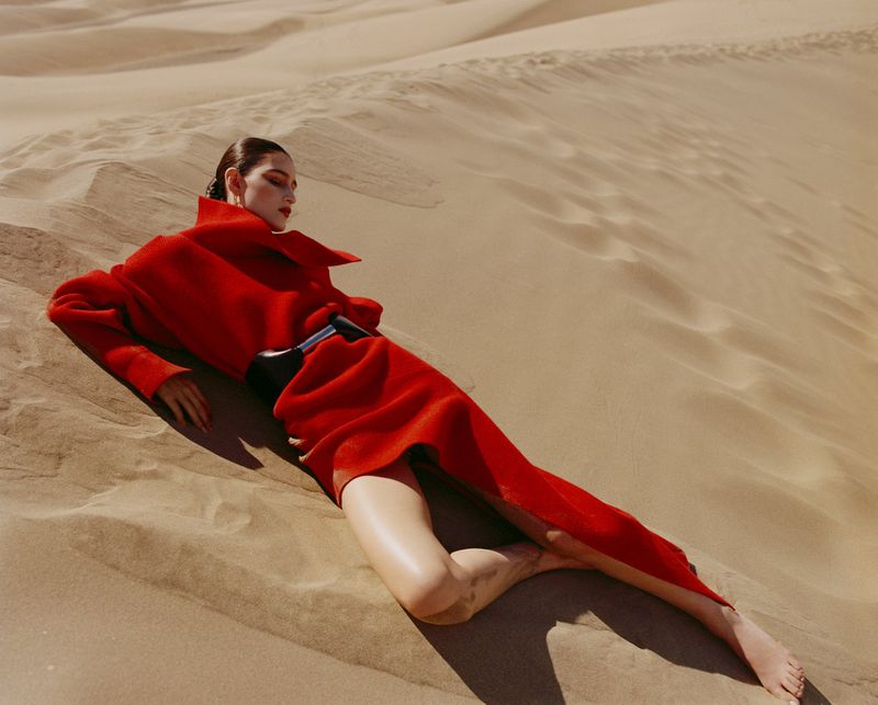 Model wearing a vibrant red coat lying on sand dunes under warm natural sunlight.