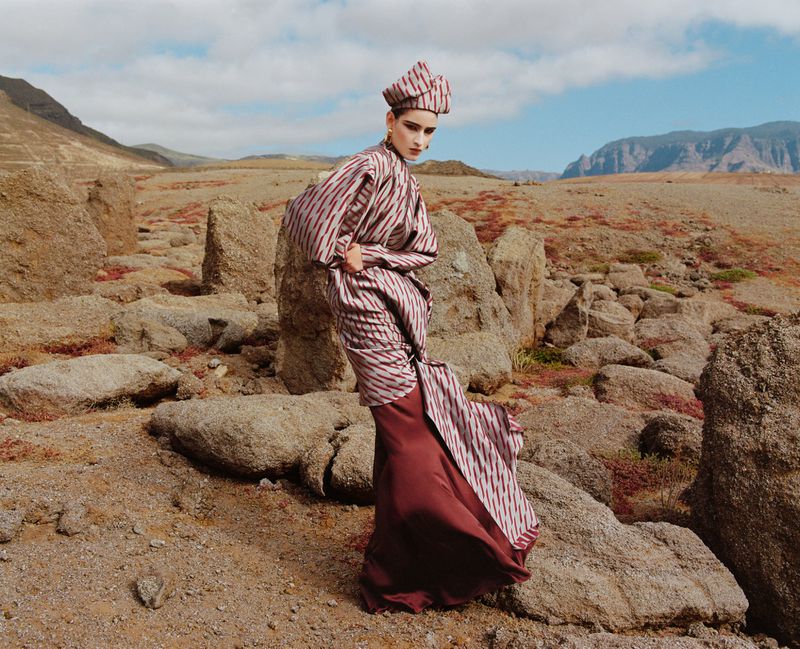 Model standing on a rocky desert landscape wearing a patterned striped tunic, matching headscarf, and a long dark red skirt.
