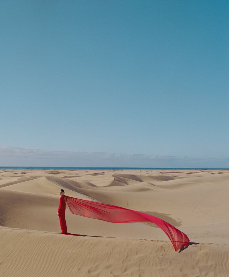 Model in a long red dress standing on a vast sand dune with a long red fabric trail blowing in the wind under a clear blue sky