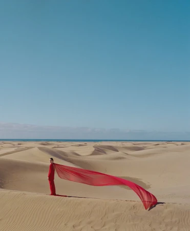 Model in a long red dress standing on a vast sand dune with a long red fabric trail blowing in the wind under a clear blue sky