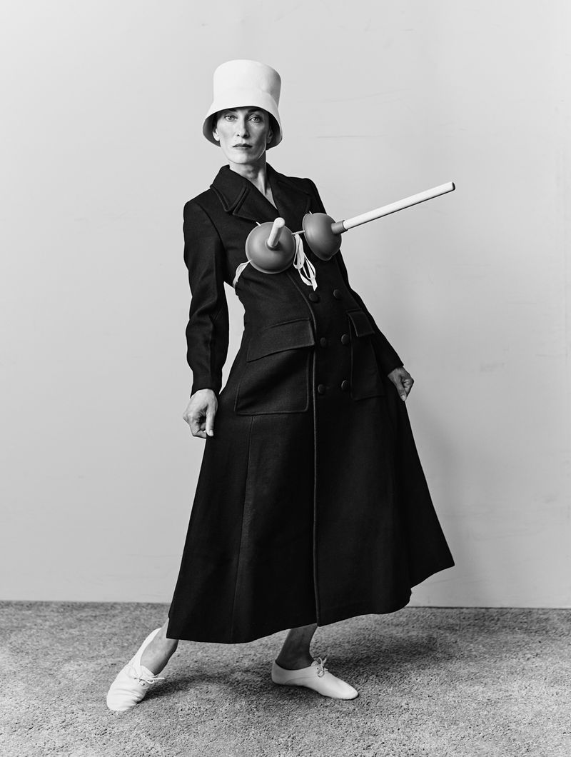 Model in a black coat with plungers attached to the chest wearing a white bucket hat and white shoes against a plain background