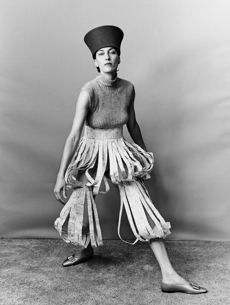 Model wearing a structured hat, sleeveless top, and fringed trousers against a grey seamless background