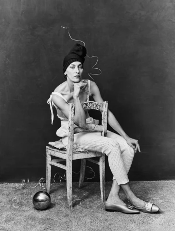 Model wearing a dark knit hat and light-colored textured trousers seated backwards on a weathered wooden chair against a dark backdrop