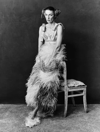 Model seated beside a wooden chair wearing a feathered skirt and ornate hairstyle against a dark background