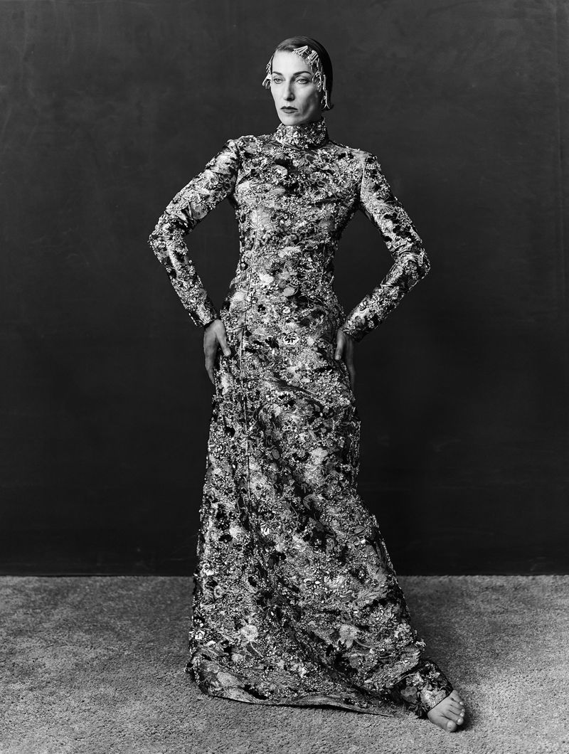 Model standing in a long patterned high-neck gown with hands on hips against a dark textured studio backdrop.
