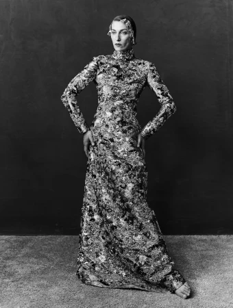 Model standing in a long patterned high-neck gown with hands on hips against a dark textured studio backdrop.