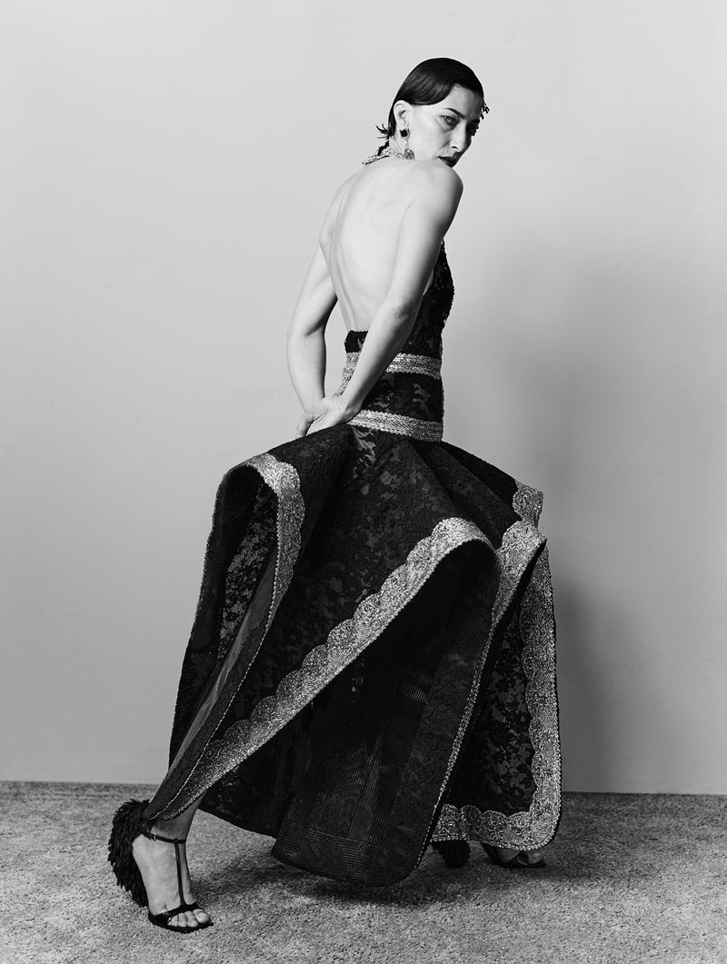 Model in a black lace structured gown looking back over her shoulder against a plain grey studio backdrop