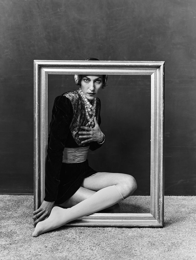 Model in dark dress holding a large ornate picture frame in a black and white fashion editorial studio portrait.