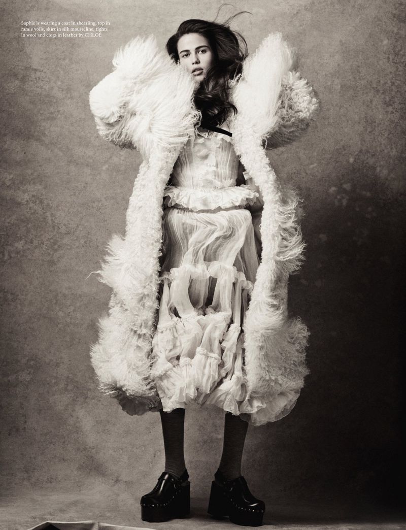 Model wearing a voluminous shearling coat and ruffled dress against a textured studio backdrop