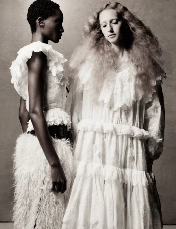 Two female models in white textured dresses posing against a muted textured backdrop in an editorial portrait.