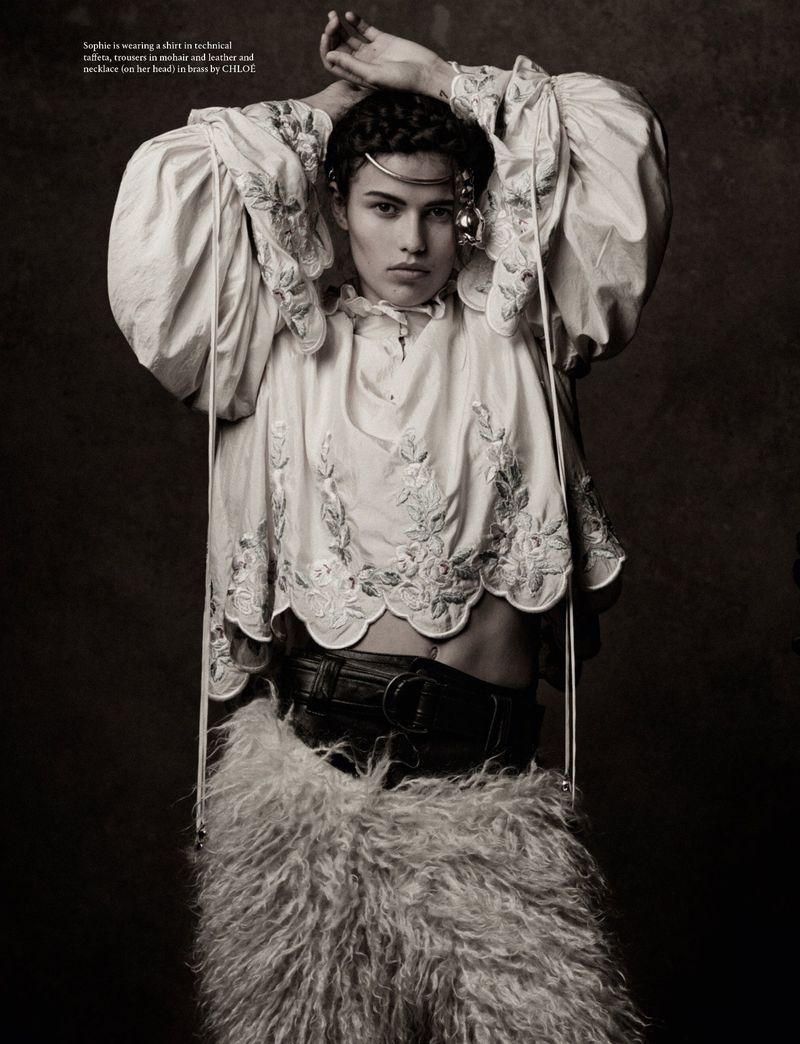 Model with braided hair wearing a white embroidered blouse and furry skirt with hands raised behind head